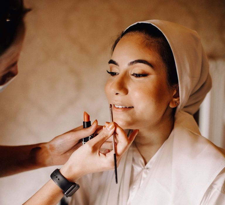 Bride gets her makeup done by a makeup artist using a peach coloured Nars lipstick and smoked wing eyeliner