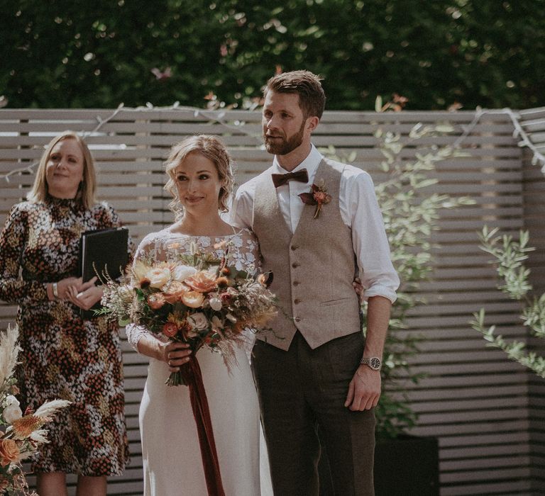 Bride in white Rime Arodaky dress with mesh floral embroidered top holding burnt orange and peach bridal bouquet with burgundy ribbon stands with groom in light brown waistcoat and bowtie during garden wedding ceremony with burnt orange wedding theme