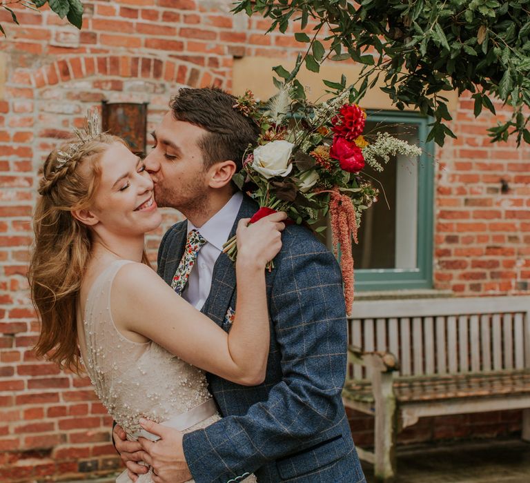 Groom in a checked suit kissing his bride with a half up half down wedding hairstyle wearing a bridal crown 