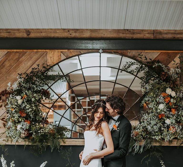 Portrait of the groom in a tuxedo embracing his bride in a strapless wedding dress and at their rustic barn wedding venue with crescent mirror and floral arrangement decor 