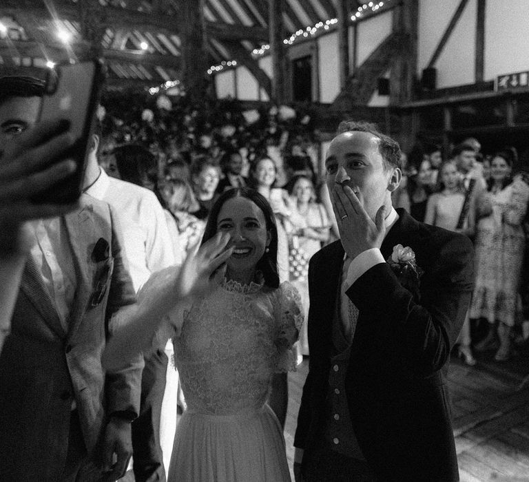  Bride in white lace puffed sleeve Daarlana wedding dress waves and groom in black morning coat blows a kiss to iPhone screen at Loseley Park wedding reception