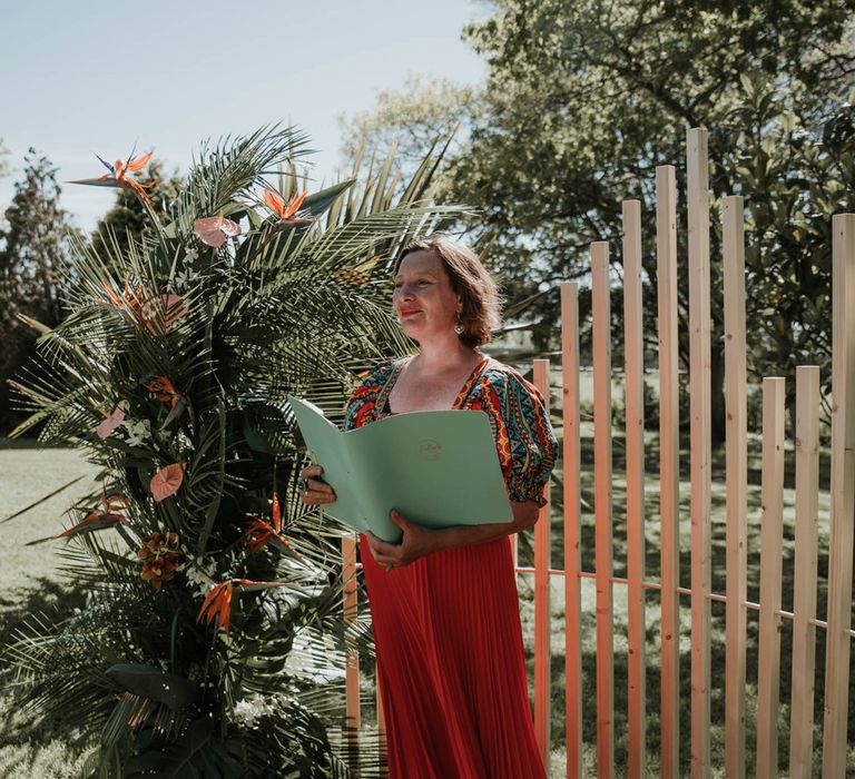 Celebrant in red dress holding green binder stands in front of white wooden poles and tropical flower installation for birds of paradise wedding inspiration