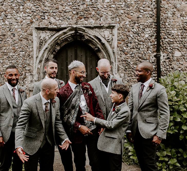 Groom in a burgundy jacquard jacket and groomsmen in grey blazer and black trouser wedding suits 