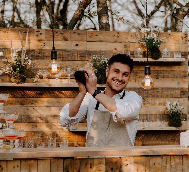 Barman shaking the cocktails behind the mobile wooden bar with copper frame and festoon lights 