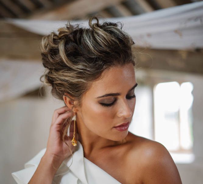 Bride in a one shoulder jumpsuit with pinned back hair wearing smokey eye makeup, highlighter on her cheeks and glossy lipstick 