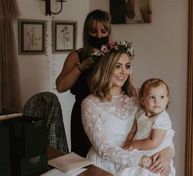 Bride on the wedding morning in a long sleeve wedding dress and pink and white flower crown with her daughter on her lap 