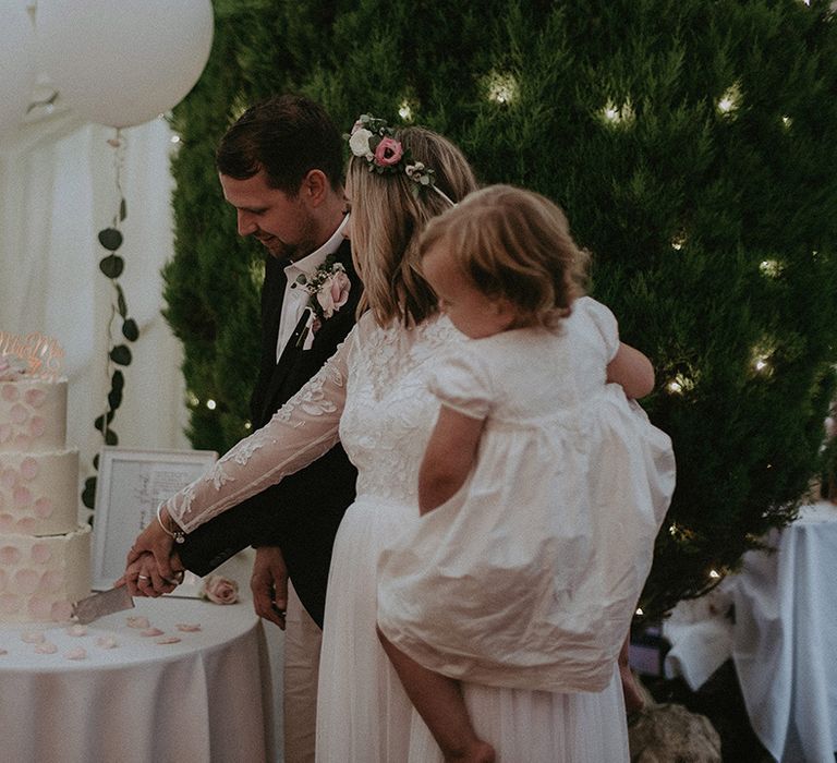 Bride and groom cutting the white and pink wedding cake with their daughter in their arms 