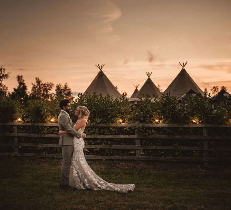 Bride in off the shoulder Enzoani wedding dress with trains stands hugging groom in grey suit as they stand by tipi at Inkersall Grange Farm wedding