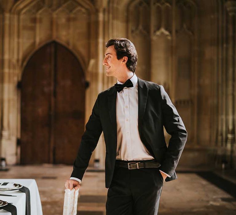 Groom in a black suit and bow tie standing at the intimate reception table decorated with black, white and gold tableware and flowers 