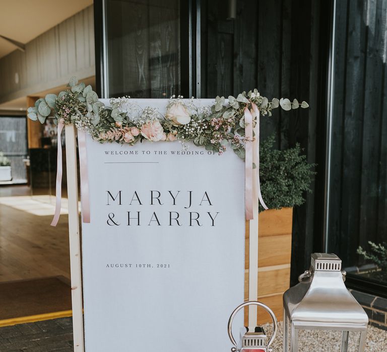 White fabric wedding welcome sign on wooden stand with floral garland, leaves and large candles in silver and glass candle holders for summer wedding at Primrose Hill Farm