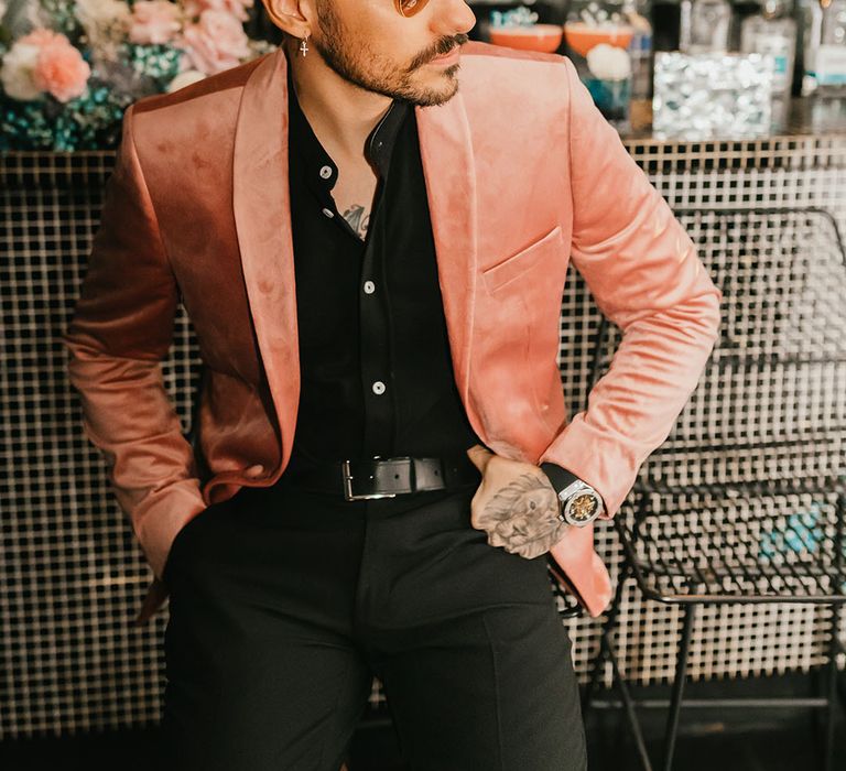 Stylish groom sitting at the bar in a black shirt, pink velvet jacket and sunglasses 