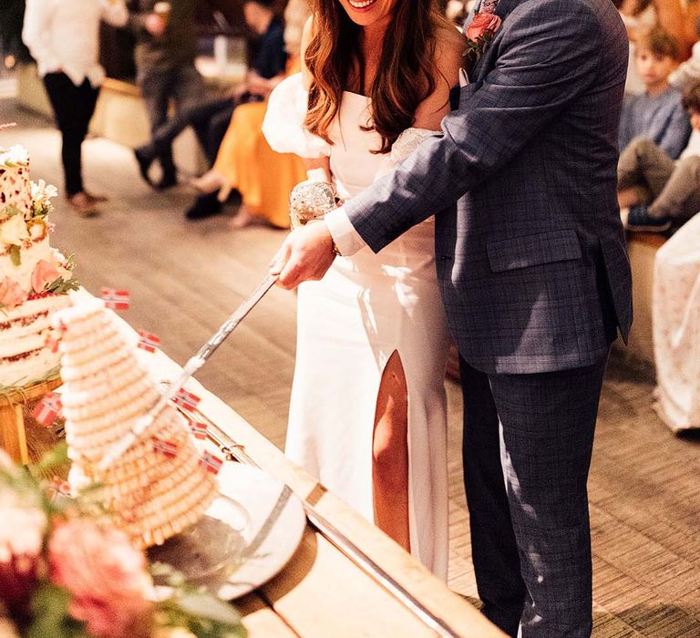 Bride in a strapless wedding dress and groom in a blue check suit cut cake with a long sword on their wedding day 