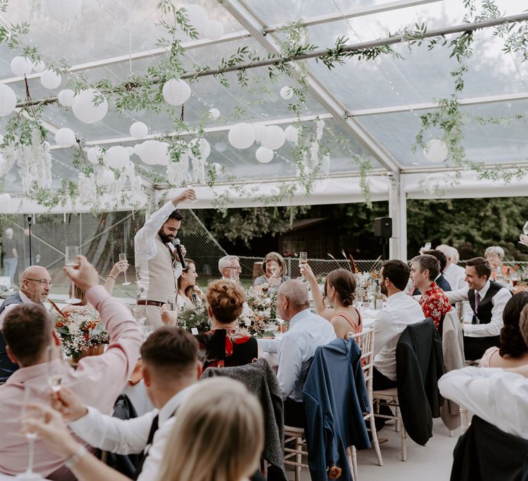 Groom in beige suit and white shirt holds dunk in the air as wedding guests raise their glasses during marquee wedding reception