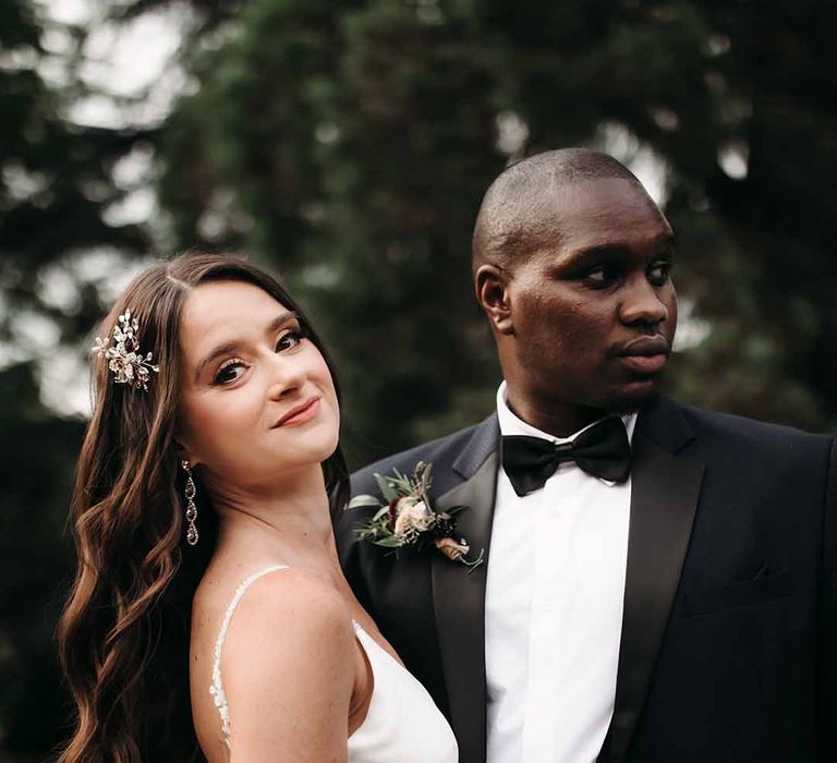 Groom in satin colour suit with black silk bow tie & Bride wearing diamond drop pearl earrings with satin button open back wedding dress 