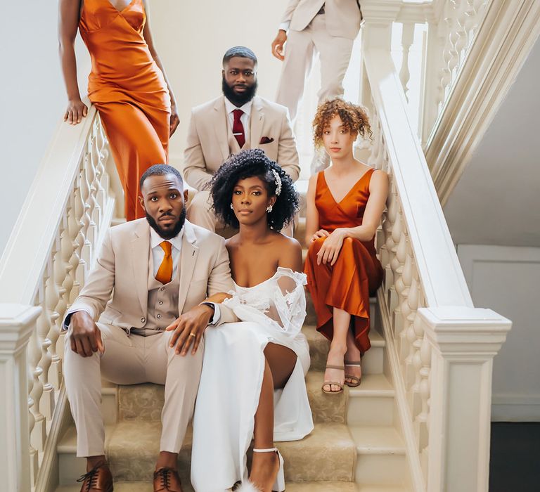 Bride with naturally curly hair in a strapless wedding dress linking arms with her groom in a beige suit on the staircase at Modern Hall with their groomsmen and bridesmaids behind them 