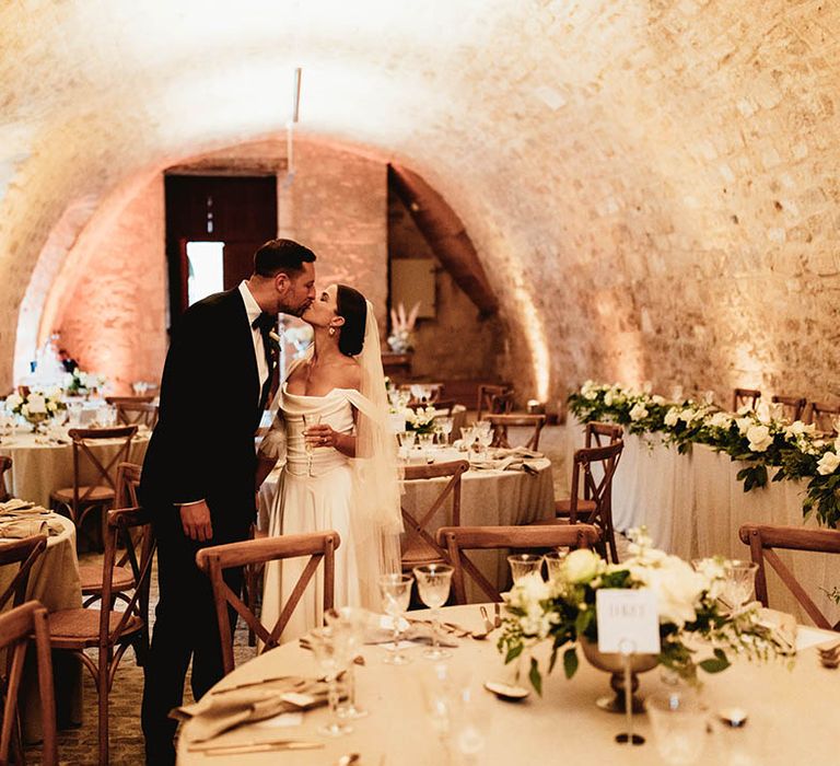 Bride & groom kiss in wedding venue for reception surrounded by classic white floral bouquets with green foliage