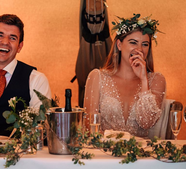 Bride in v neck embroidered Julie Vino wedding dress and boho flower crown sits laughing with groom in red tie and waistcoat as they sit at top table for tipi wedding reception