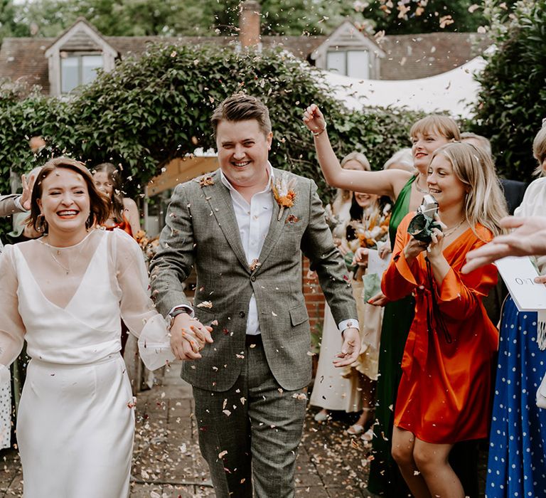 Confetti moment with bride in a long sleeve wedding dress holding an orange dried flower bouquet and groom in a grey check suit 