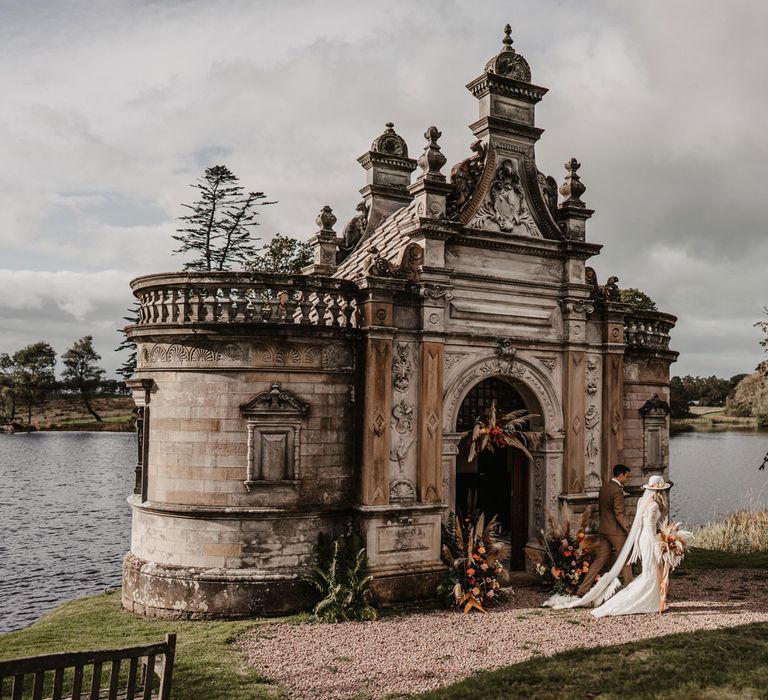 Bride in boho wedding dress and hat walks with groom in brown tweed suit next to The Bathing House at Kinmount House, Scotland 