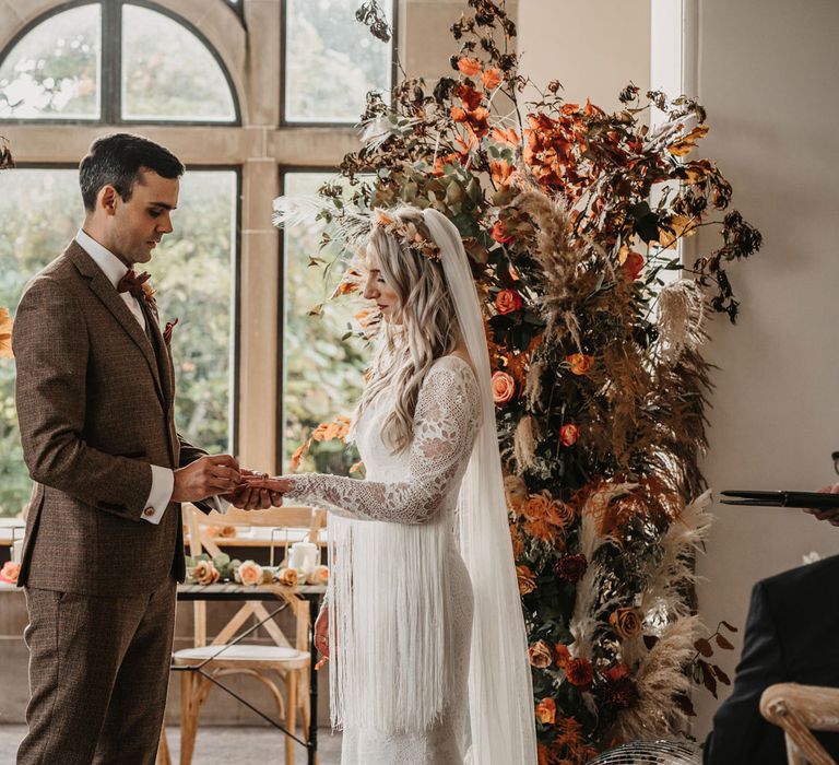 Groom in brown three piece tweed suit puts ring on finger of bride in two piece lace wedding dress with tassel sleeves and veil during intimate wedding ceremony at Kinmount House with orange and green boho florals