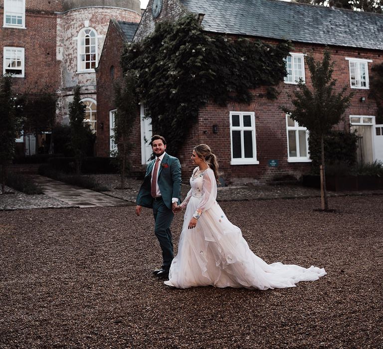 Groom in blue suit and pink tie walks holding hands with bride in bespoke Victoria Sanders wedding dress with train in the grounds of Iscoyd Park