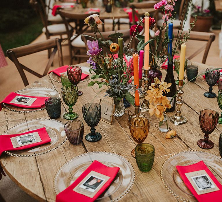 Round wedding reception table with clear glass charger plates, pink napkins, polaroid name place cards and colourful candles, vases and flower stems centrepiece decor 