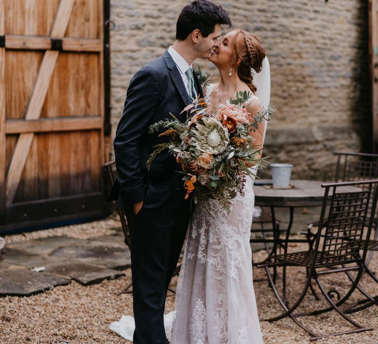 Bride in lace and tulle wedding dress with veil holding large bridal bouquet kisses groom in dark suit in barn courtyard 