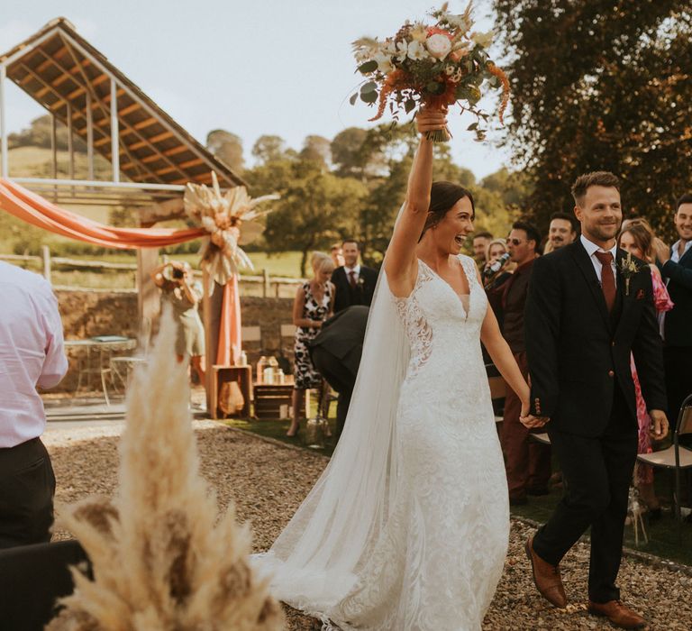 Bride in v neck lace wedding dress and veil holdings up mixed bridal bouquet as she walks back down the outdoor ceremony aisle with groom in dark suit and burgundy tie