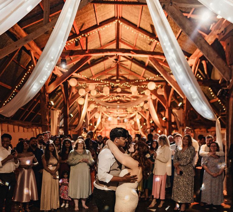 Bride & groom lean in to kiss in front of wedding party as barn venue lights up behind them complete with hanging lanterns 