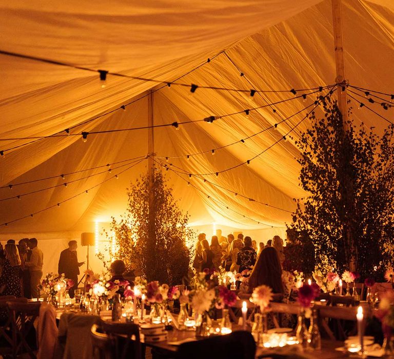 Interior of marquee wedding reception with colourful florals and foliage lit for evening wedding reception