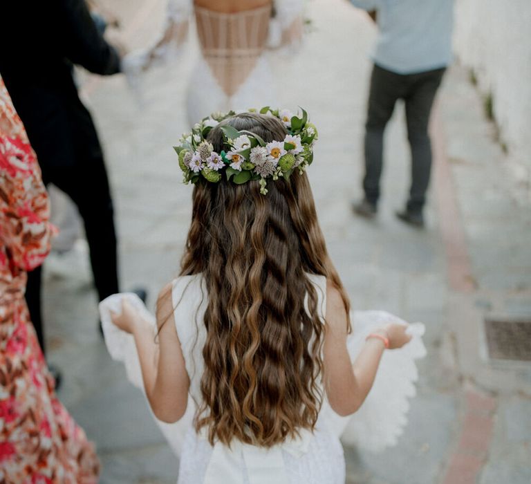 Flower girl holds train of wedding gown whilst wearing flower crown and her long brown hair in loose waves 