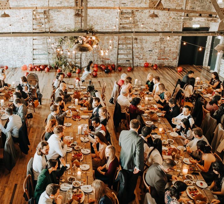 Wedding guests sit at long wooden table in industrial style wedding reception room with beams and exposed brick 