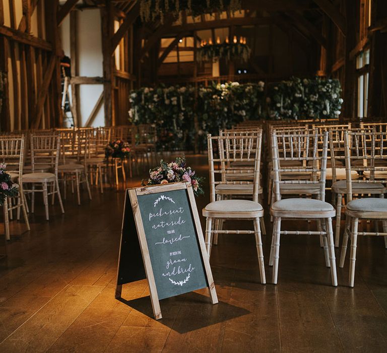 Rustic wedding venue with light wooden and cream seats with flower displays and chalk notice board reading 'Pick a seat, not a side. You are loved by both the bride and groom.'