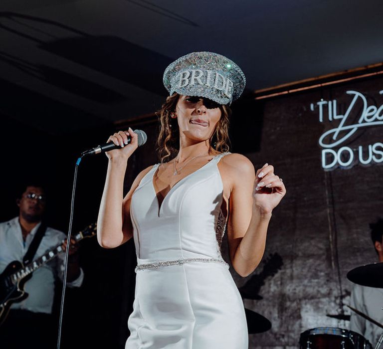 Bride in a V neck wedding dress and jewelled Bride hat signing on stage with a  'Til death Do Us Party' Neon wedding sign behind her