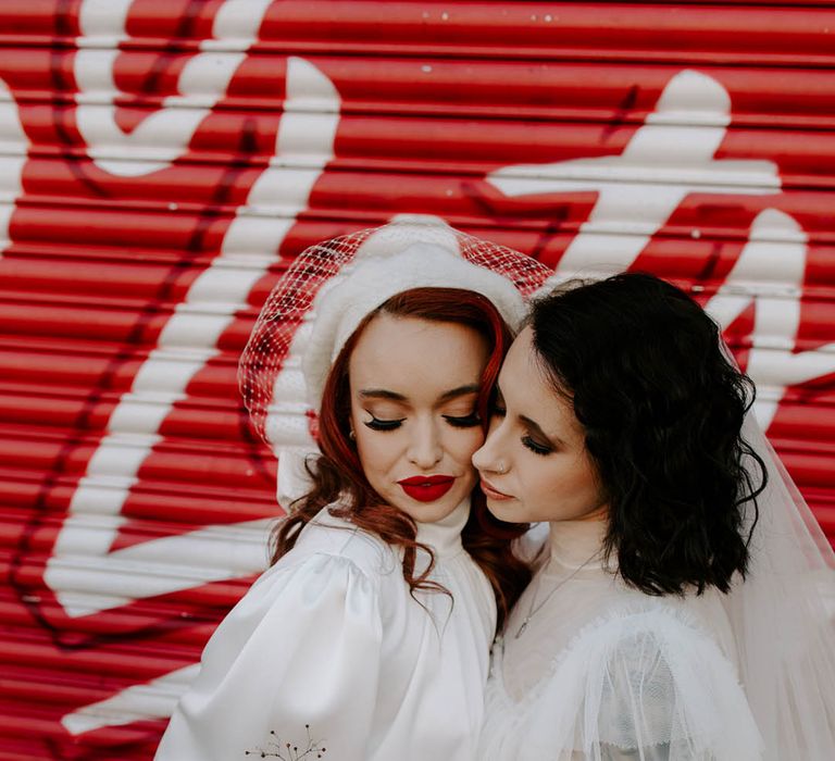 Two brides embracing in tulle and satin short wedding dresses with high necks holding a red and pink romantic wedding bouquet 