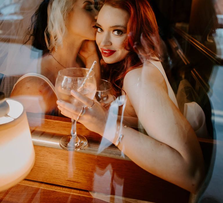 Bride with two tones hair kissing her bride with red hair as she sips gin from a balloon glass 