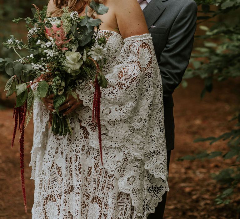 Groom in grey suit and pink tie hugs the bride in lace wedding dress and flower crown for woodland wedding