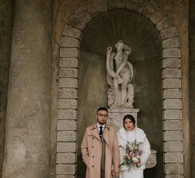 Bride holding mixed neutral bouquet in white fur cropped coat under statue 