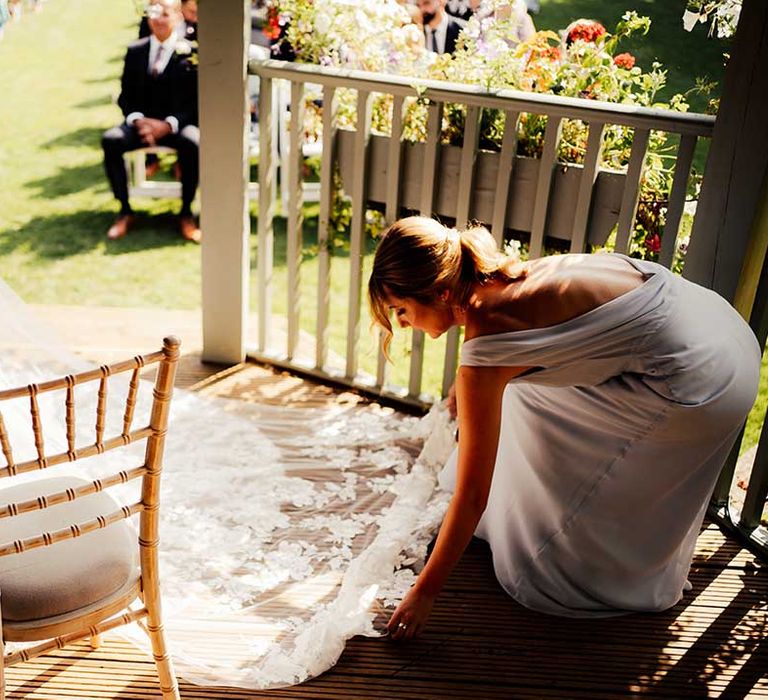 Bridesmaid in grey low back dress lays out the bride's lace floral veil out behind the bride