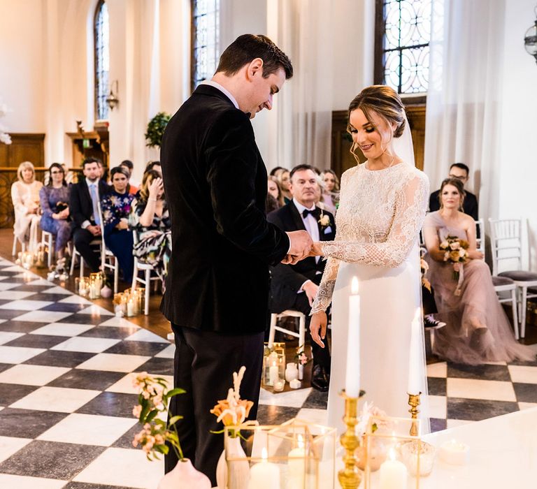 Groom in black tie puts the ring on the bride's finger wearing lace top long sleeve wedding dress