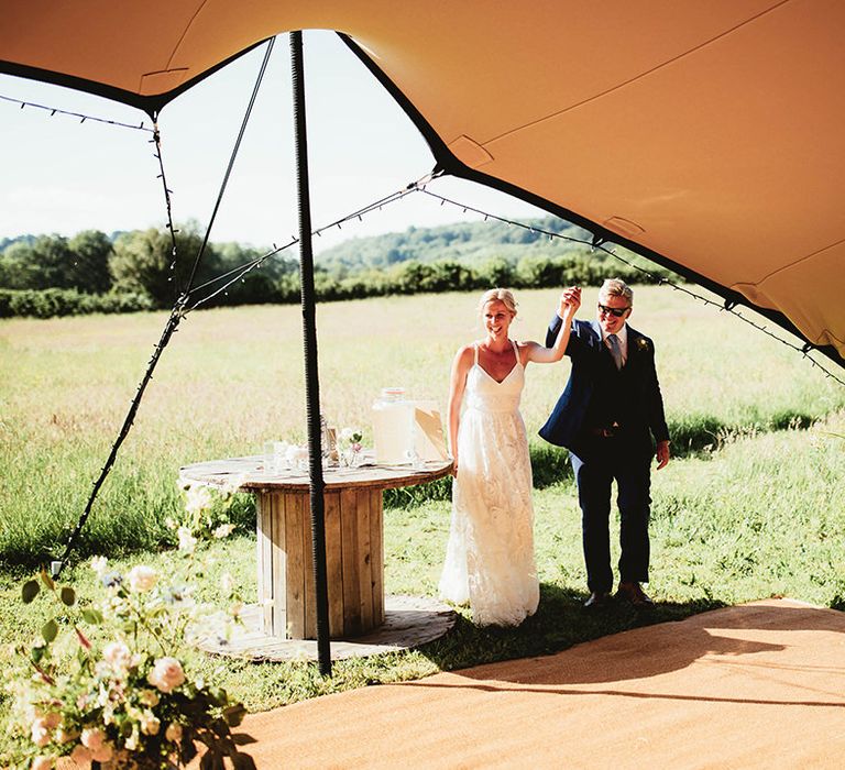 Bride and groom enter stretch tent for reception with fairy lights and vintage milk churns full of flowers 