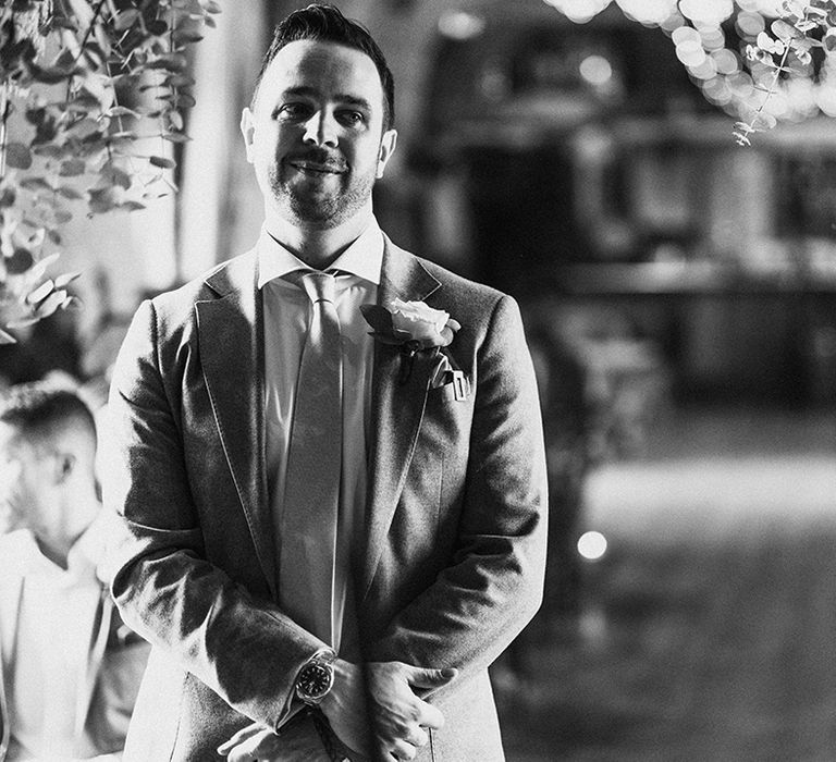 Groom waits at the altar for the bride wearing grey suit with white rose buttonhole