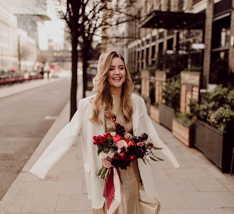 Bride in gold wedding dress and small handbag with white blazer cover up and pink, red and purple wedding bouquet 