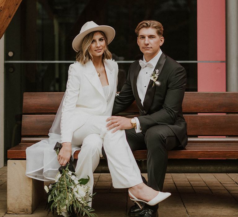 Groom sits in black tie with his hand on the bride's knee as she wears white trouser wedding suit and white slingback heels