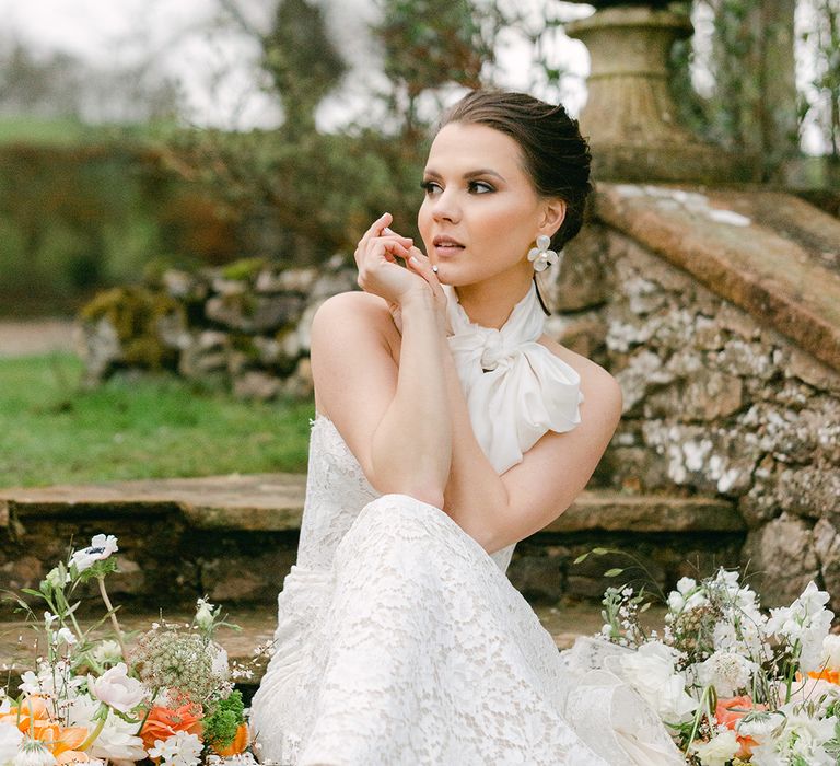 Bride with a pinned wedding up do in a strapless lace wedding dress with bow neck scarf sitting on the steps at Holesfoot