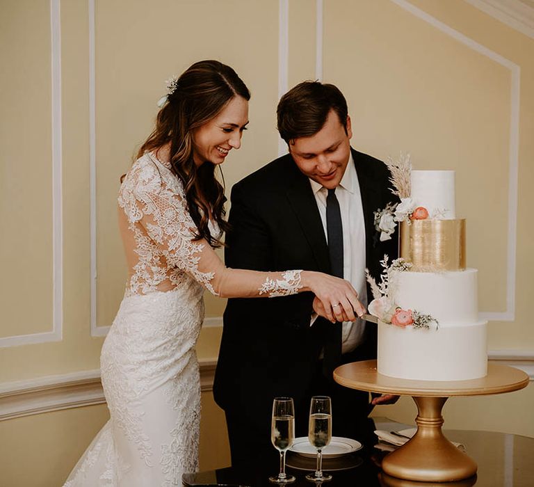 Bride and groom cut their four tier white and gold wedding cake decorated with flowers