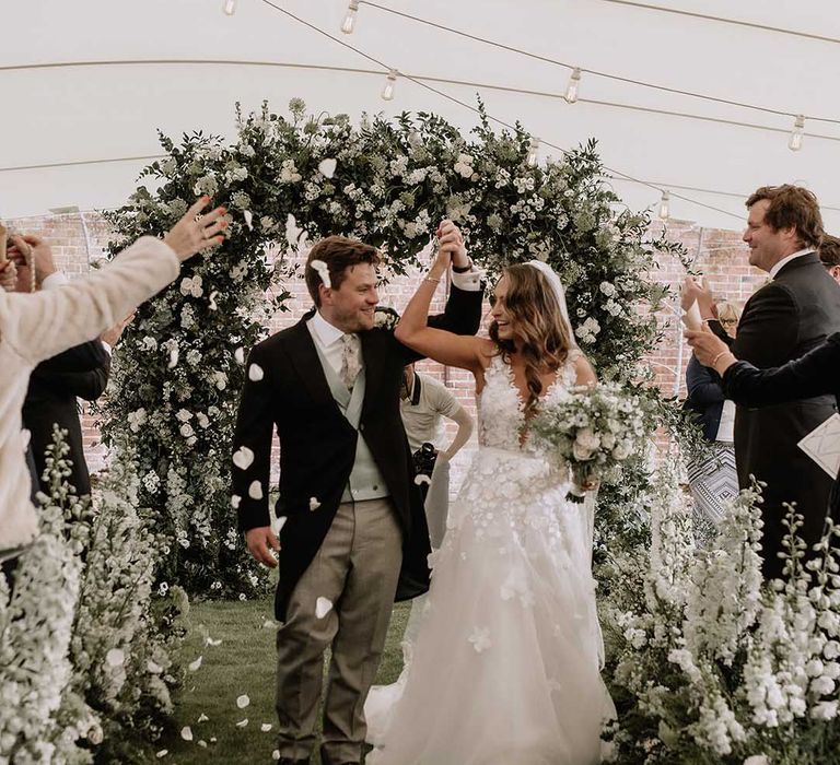 Bride and groom lift their intertwined hands as they walk back down the aisle to confetti as a married couple