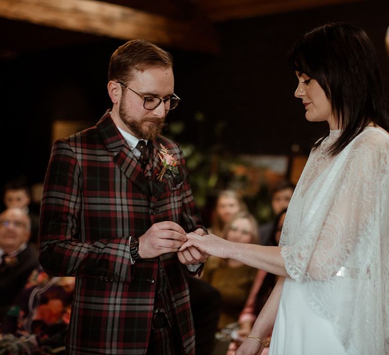 Groom places ring on his bride's finger who wears lace cape on her wedding day