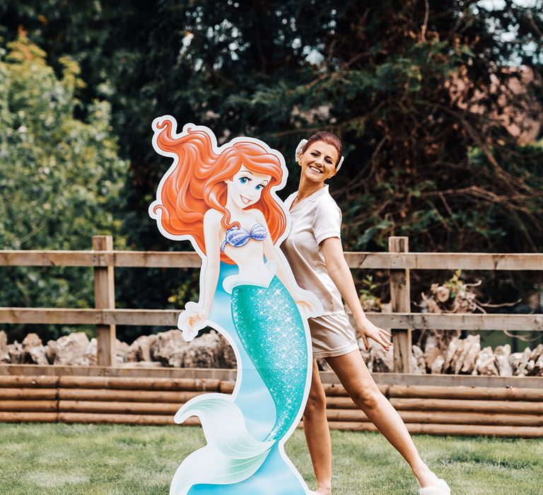 Bride in satin short pyjama set with red hair and white hair bow posing next to cardboard cutout of princess Ariel 