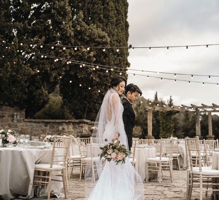 Bride wearing pearl embellished veil carries pastel floral bouquet and walks alongside groom through outdoor reception in Barcelona